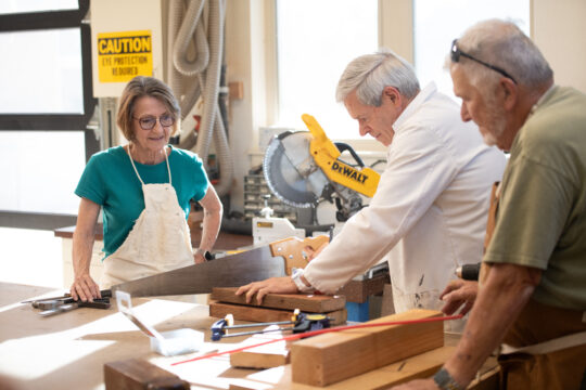 Three Paradise Valley Estates residents work in the woodworking shop. One resident uses a saw to cut a piece of wood.